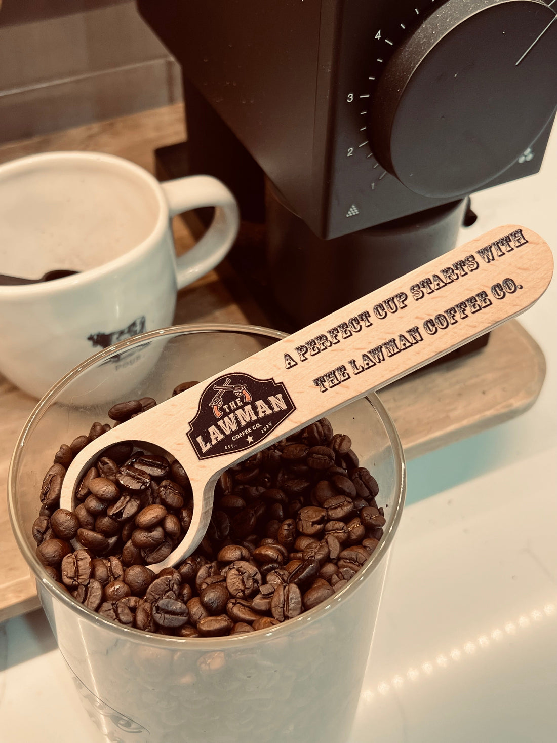 Lawman Coffee Co. branded wooden coffee scoop with whole coffee beans beside a coffee grinder and mug.